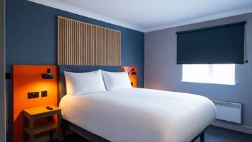 A hotel room with a bed, a nightstand, a wall-mounted lamp, a window with a blind, and a radiator.