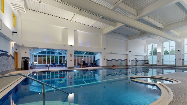 An indoor swimming pool with clear blue water, a tiled deck surrounding the pool, and a covered seating area with chairs and tables. - Village Hotel Bournemouth (Bournemouth)