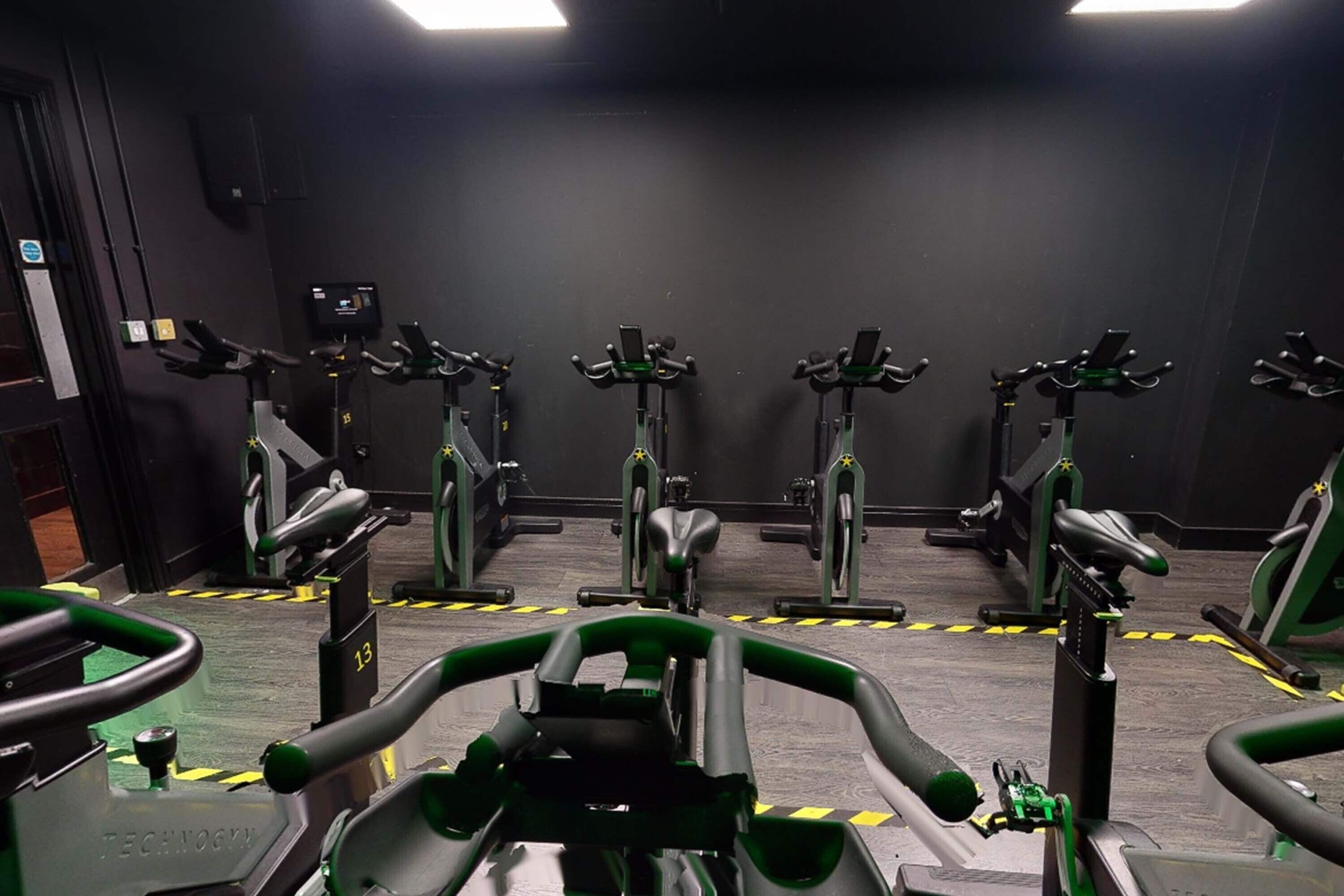 A gym with multiple stationary bikes arranged in rows, a control panel, and a wall-mounted screen.