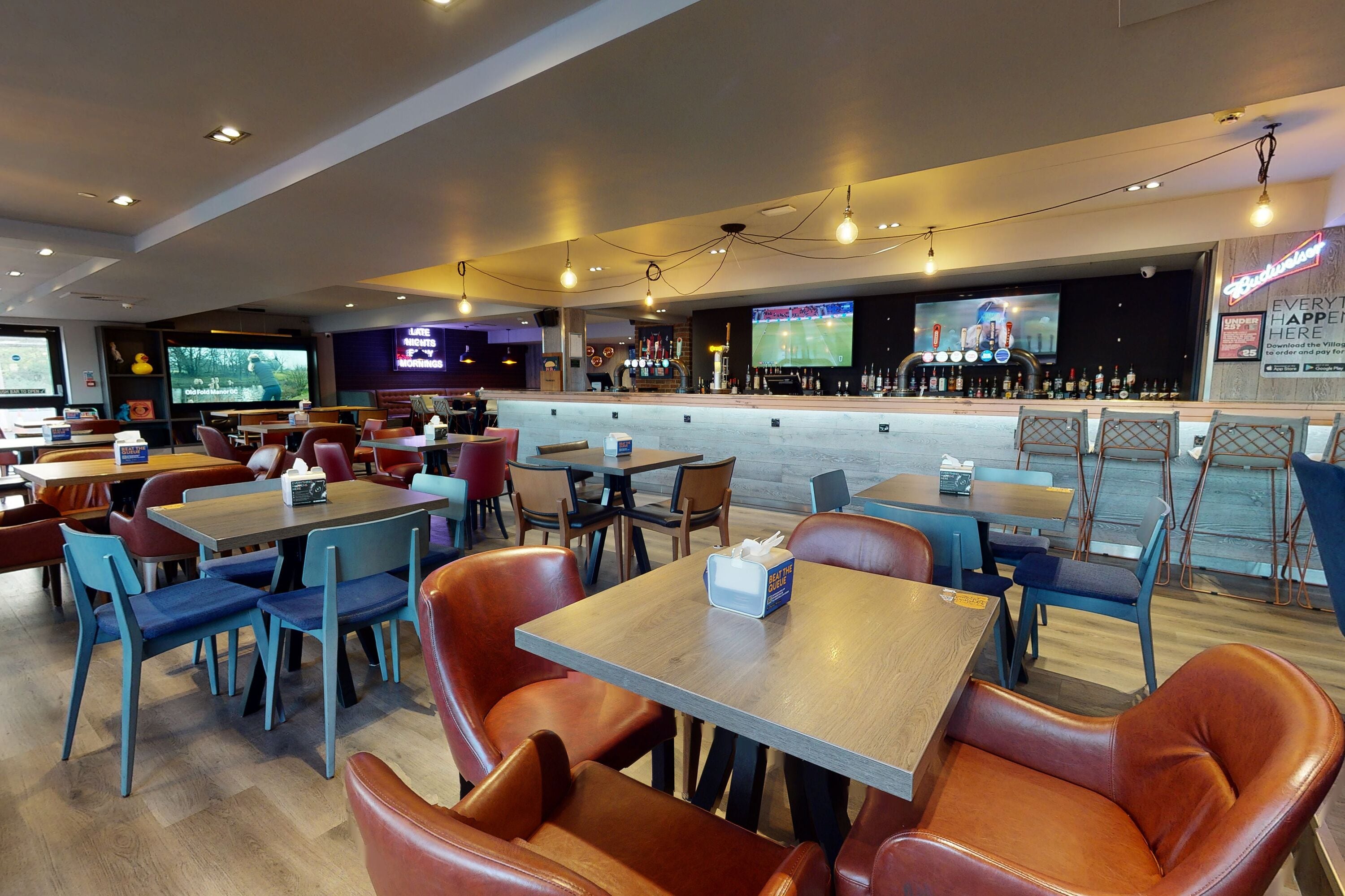 A modern restaurant with a bar area, multiple seating options including booths and tables, and a large screen displaying sports events.