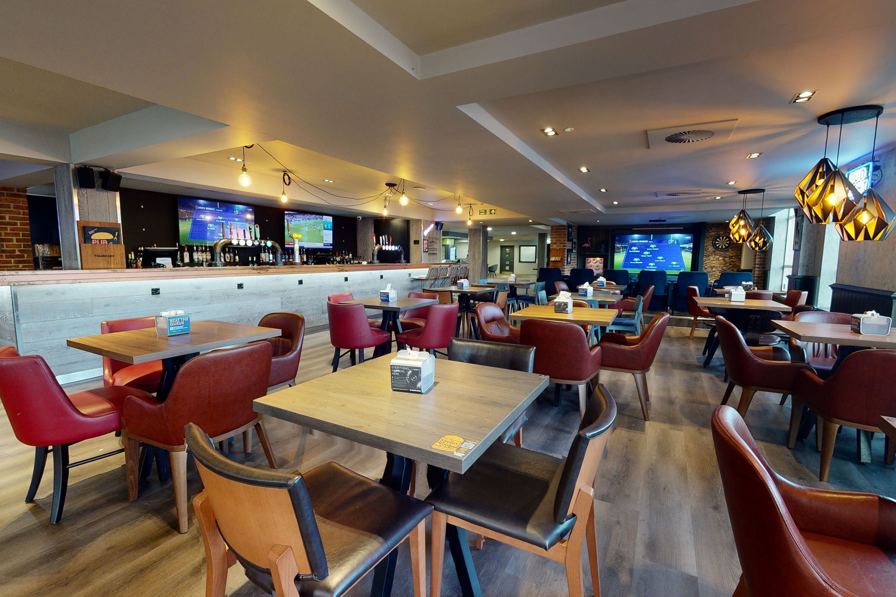 En moderne restaurant med flere borde og røde stole, et barområde og sports-tv på væggene.