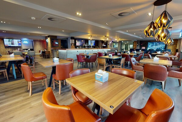 Un restaurant moderne avec des tables en bois et des chaises orange, un espace bar et plusieurs écrans diffusant du contenu.