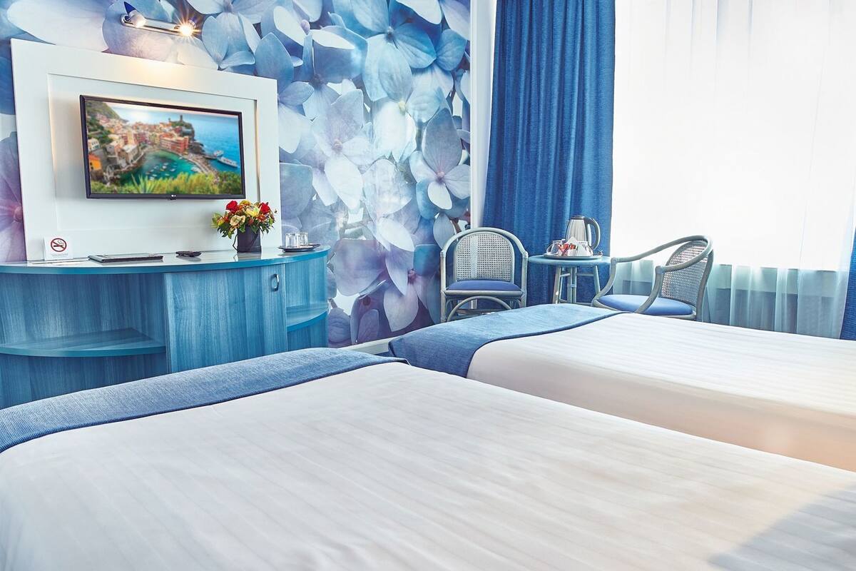 Une chambre d’hôtel avec deux lits, un bureau avec une télévision, une chaise et un mur orné d’un motif floral.