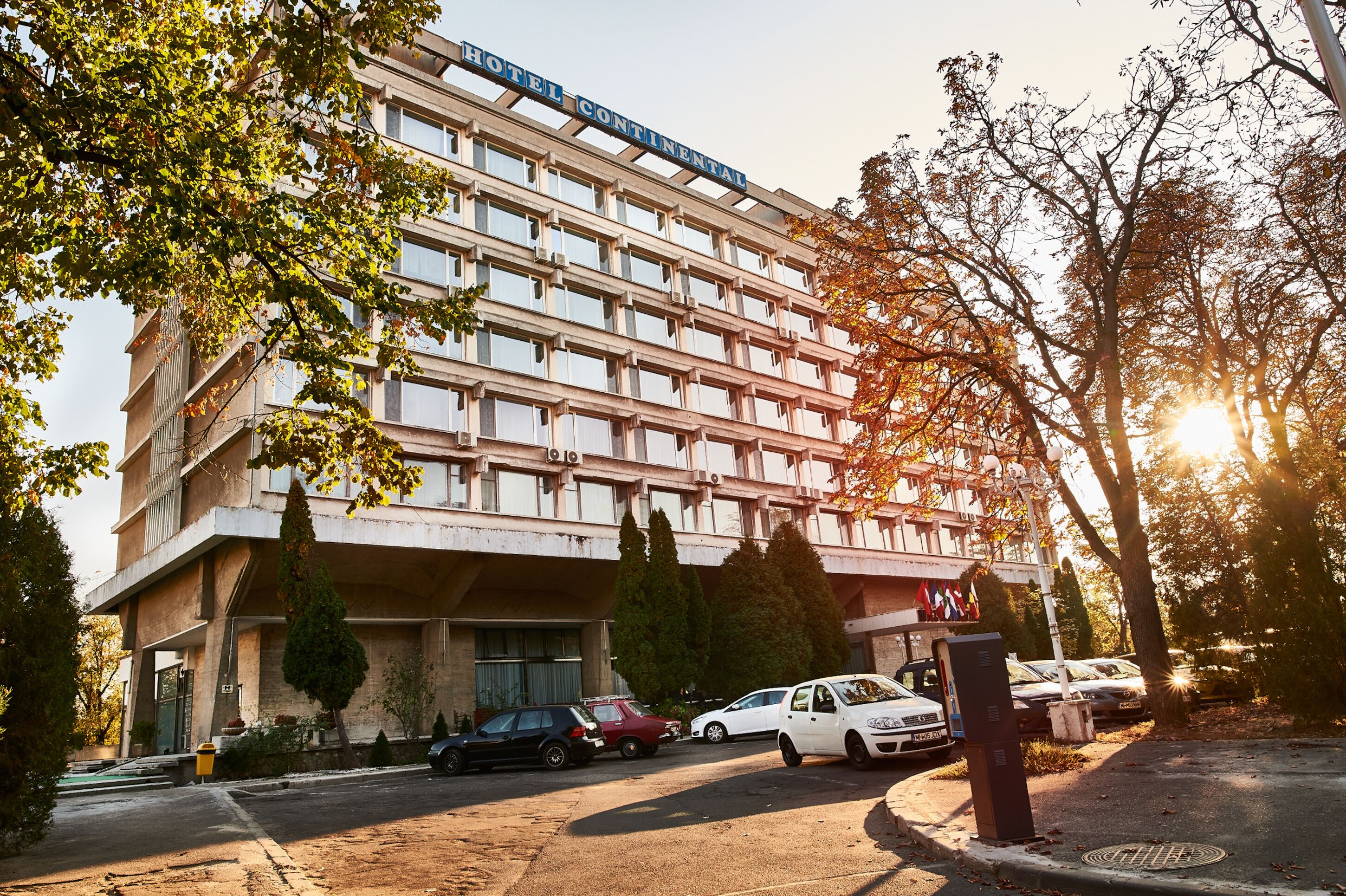Ein mehrstöckiges Hotel mit schlichter Fassade, mehreren Balkonen und einem Parkplatz mit Autos.