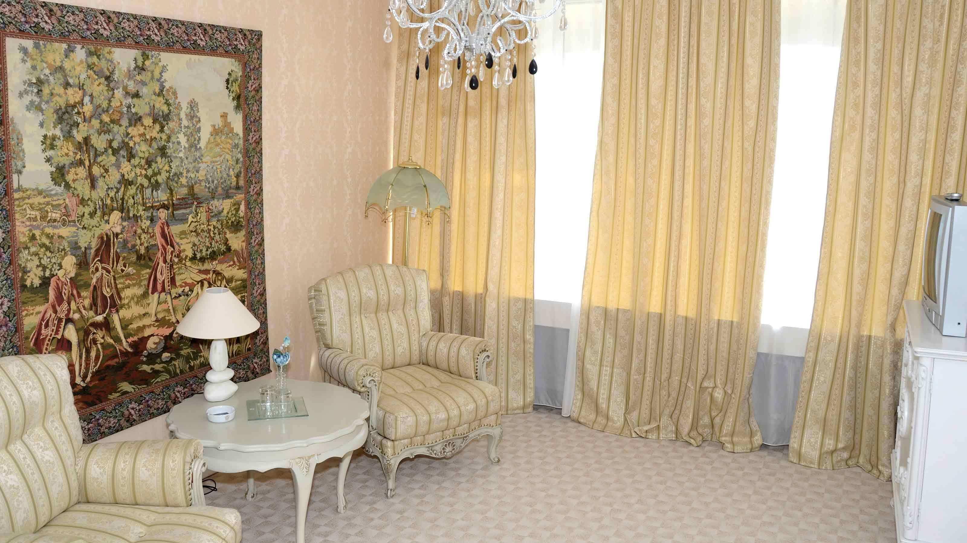 Een kamer met een groot bloementapijt, een kroonluchter, gestreepte fauteuils, een bijzettafeltje met een lamp en een televisie op een kast.