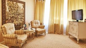 A room with a large framed painting, two upholstered chairs, a small round table, a television on a cabinet, and a patterned carpet.