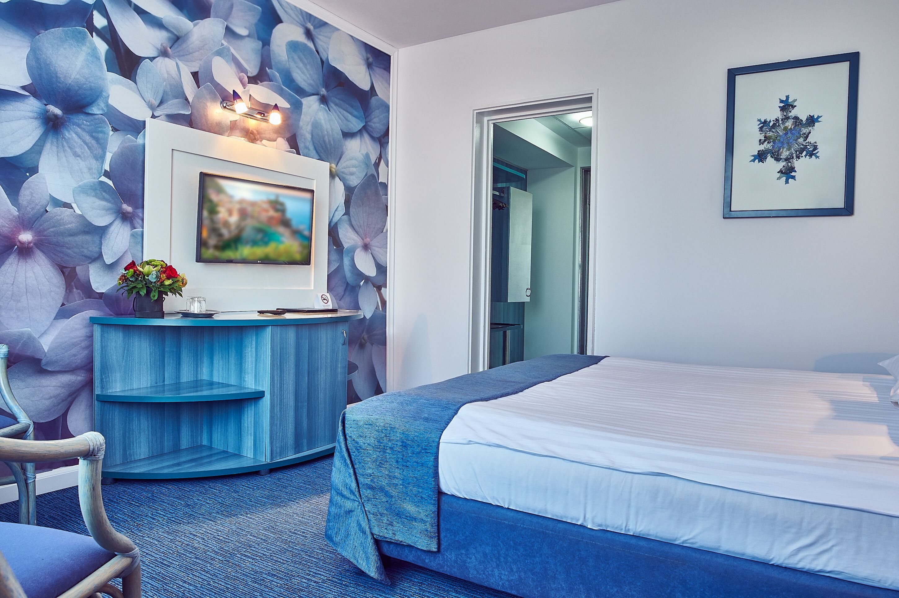 Une chambre d’hôtel avec un lit, un téléviseur posé sur une console bleue, un vase avec des fleurs et un tableau encadré accroché au mur.