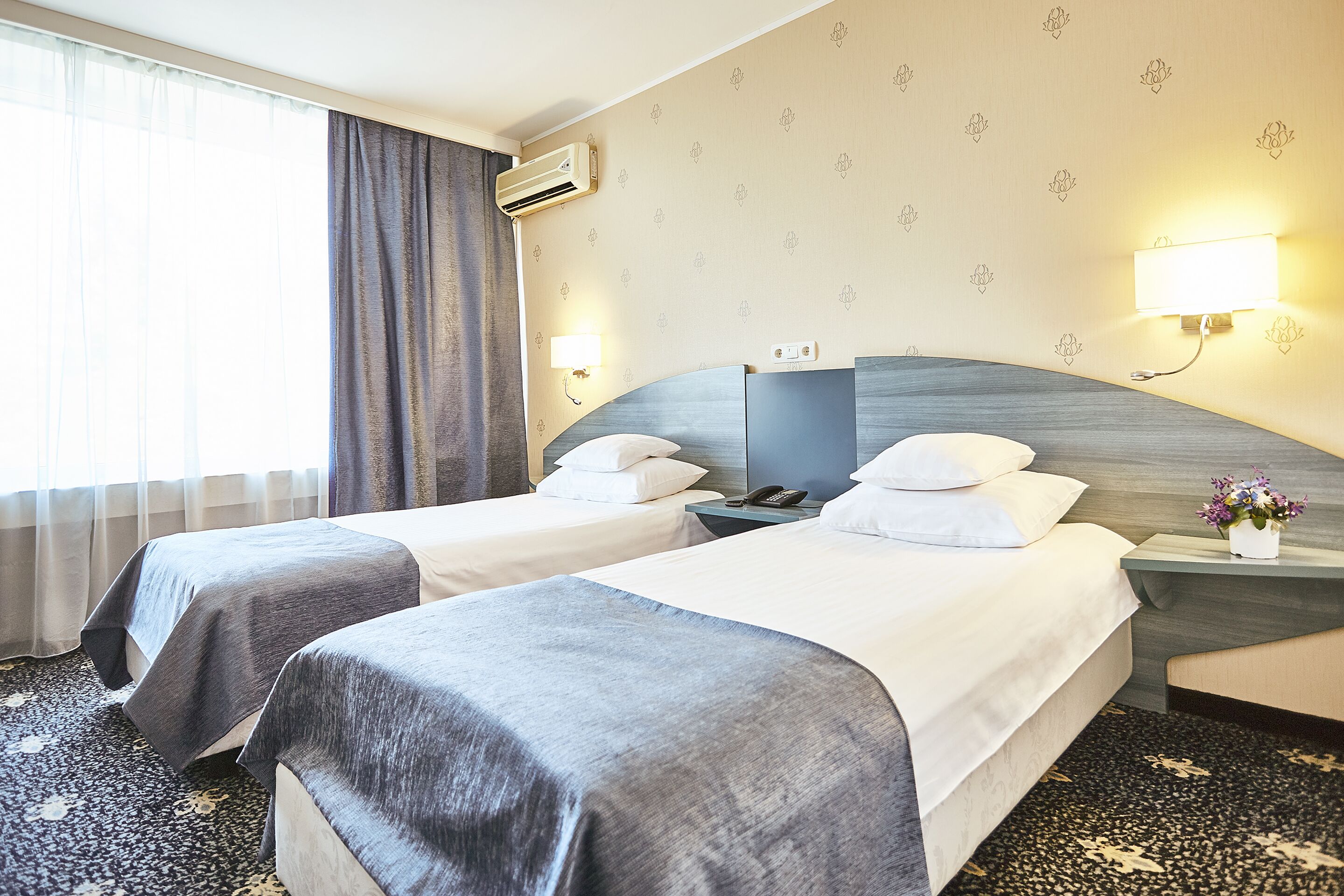 A hotel room with two beds, a nightstand with a lamp, an air conditioner, and a floral-patterned wall.