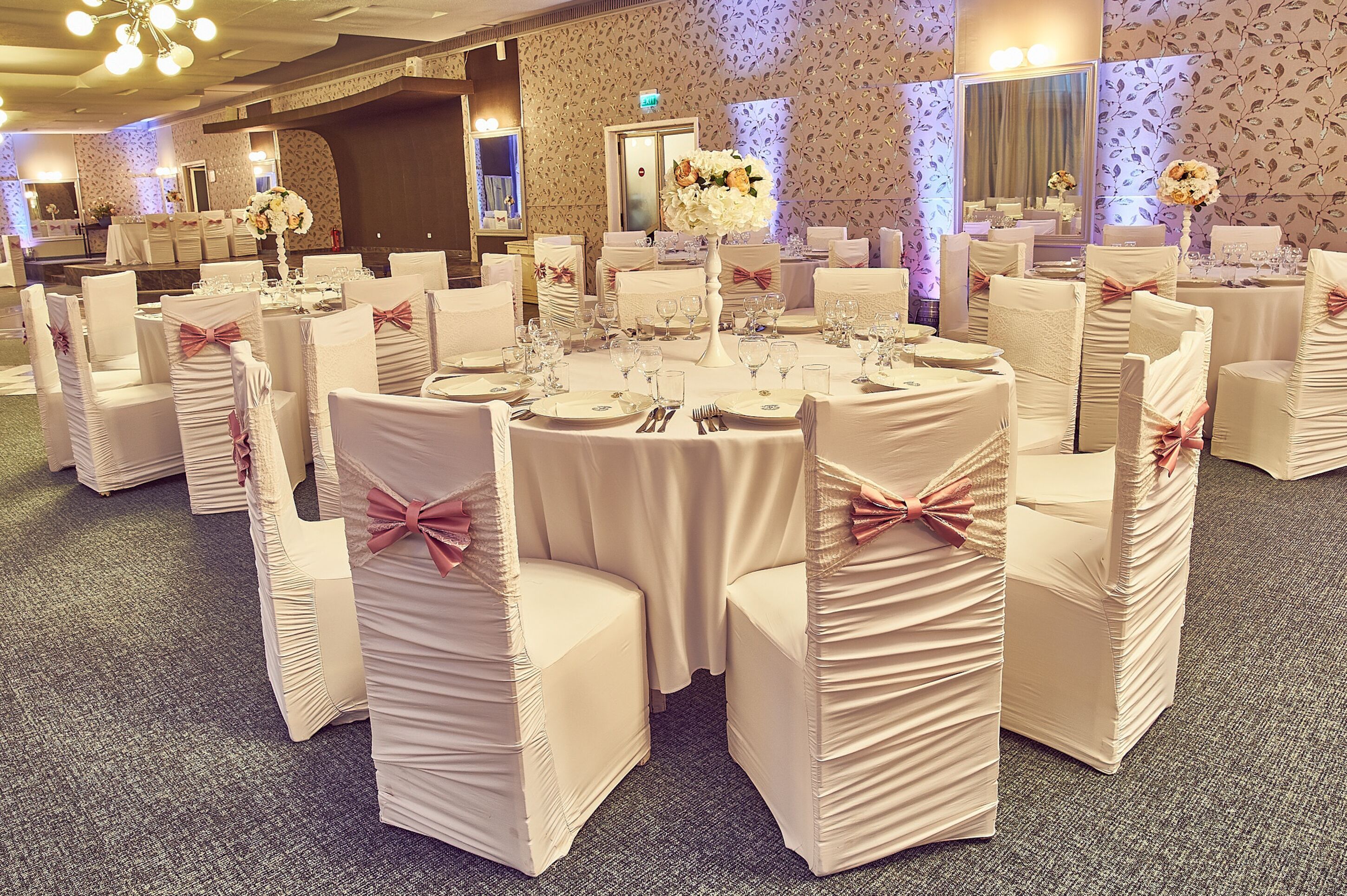 A banquet hall with round tables, white chairs with pink bows, and floral centerpieces.
