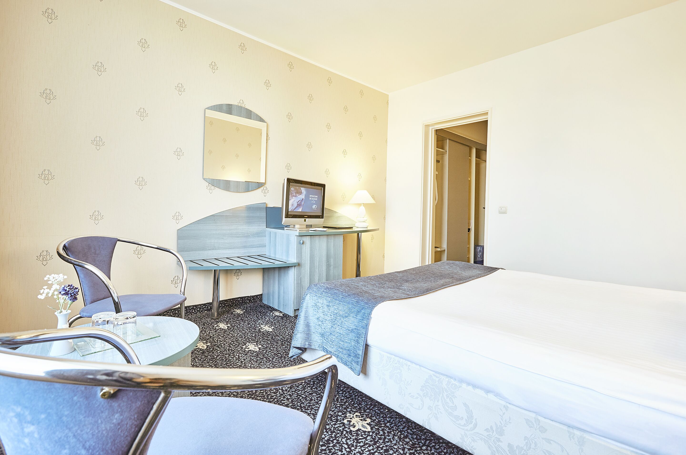 A hotel room with a bed, a desk with a computer, a chair, and a small table with a vase of flowers.