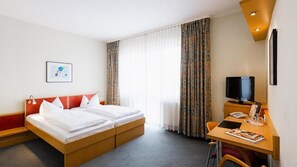 Une chambre d’hôtel avec un lit, un bureau et une chaise, une télévision et une fenêtre avec des rideaux.