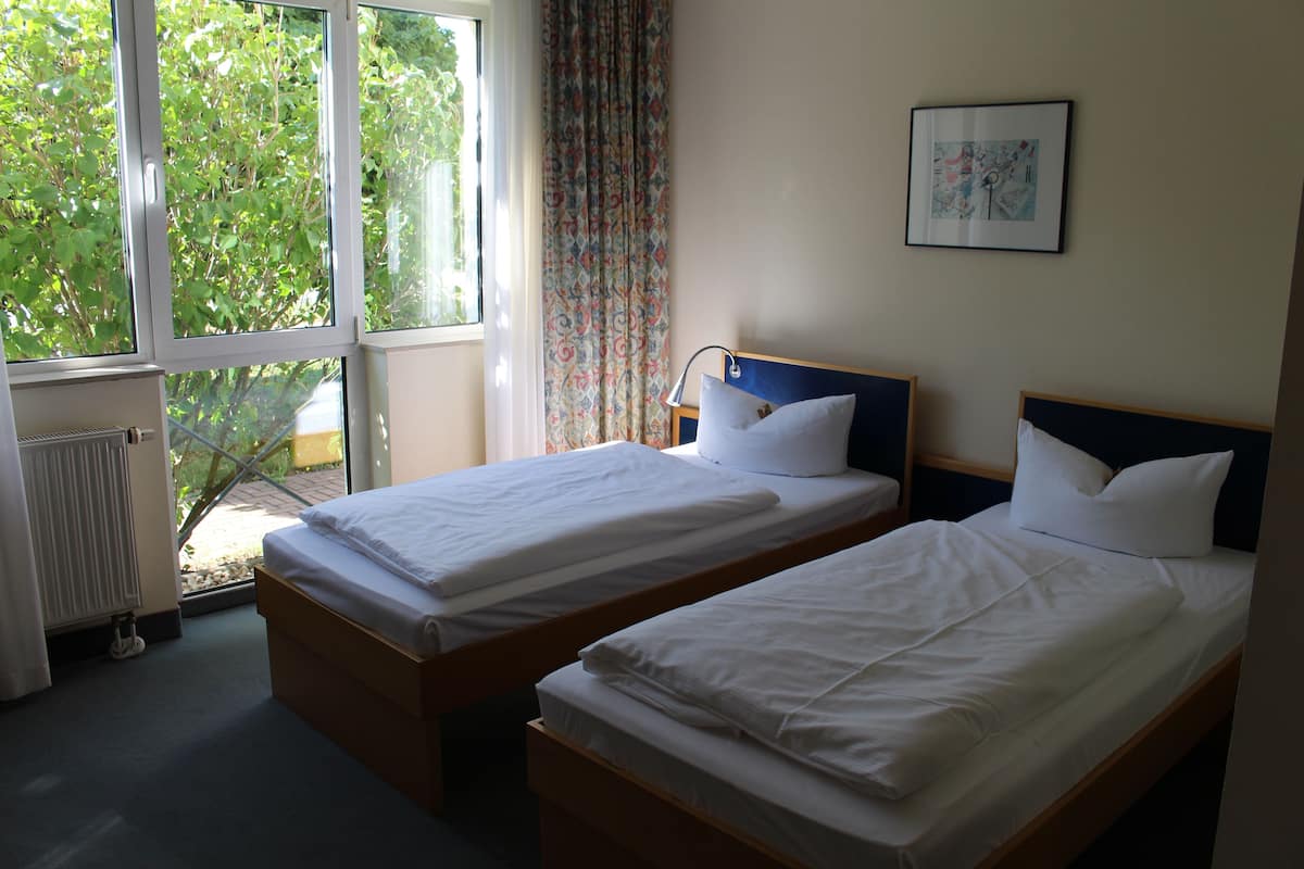 A room with two single beds, a window with a view of greenery, a radiator, and a framed picture on the wall.