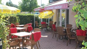 Un espace café en plein air avec des tables et des chaises en osier, certaines recouvertes de nappes rouges, et des parasols jaunes.