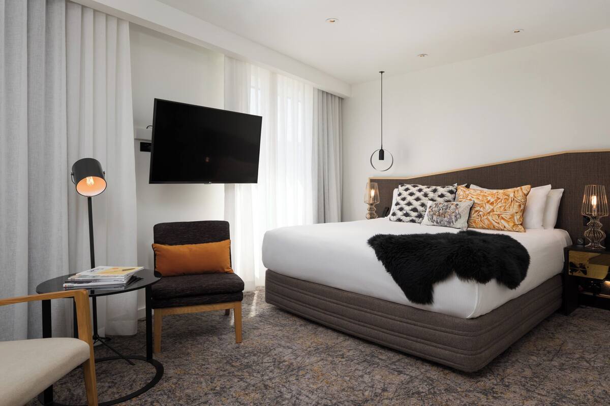 A hotel room with a large bed, a flat-screen TV mounted on the wall, a desk with a lamp, and a chair.