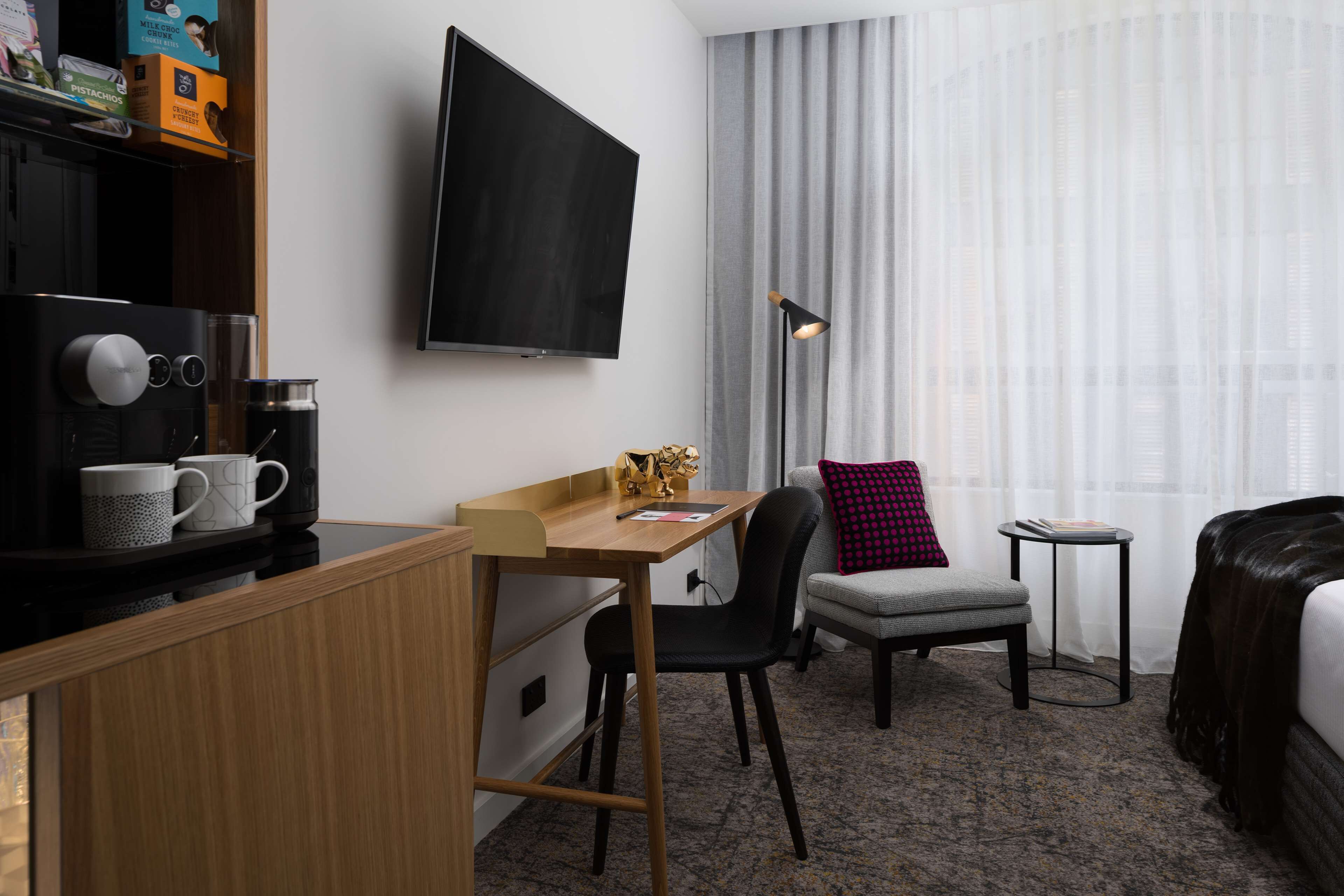 Habitación de hotel con un escritorio de madera, televisor montado en la pared, un sillón gris, una mesita auxiliar y una cama con cubrecama oscuro.