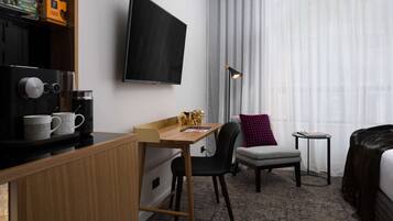 Habitación de hotel con un escritorio de madera, televisor montado en la pared, un sillón gris, una mesita auxiliar y una cama con cubrecama oscuro.