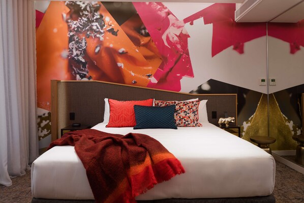 Una habitación de hotel moderna con una cama grande, varios almohadones, una manta roja y un mural en la pared.