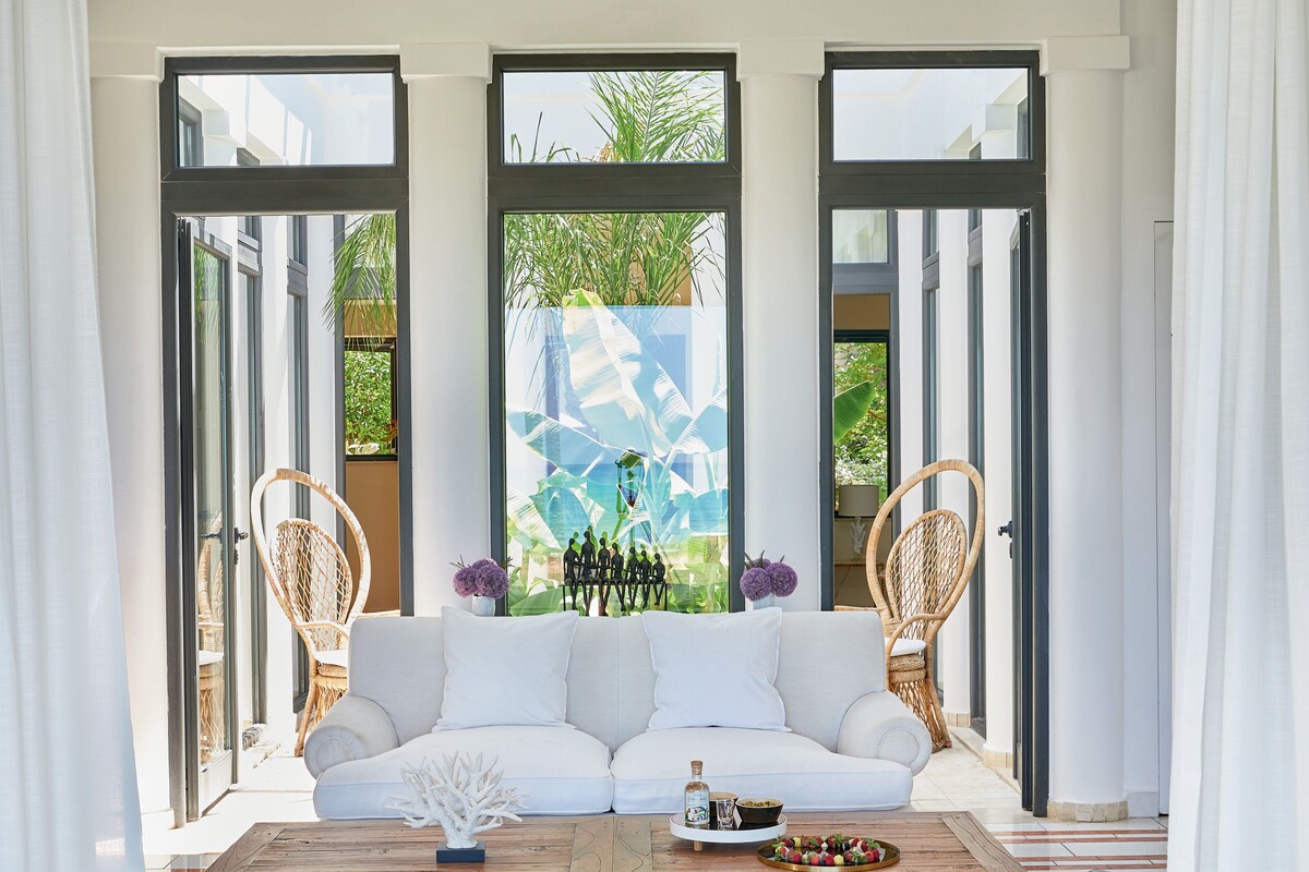 A modern living room with a white sofa, two wicker chairs, a wooden coffee table, and a glass wall with tropical plants.