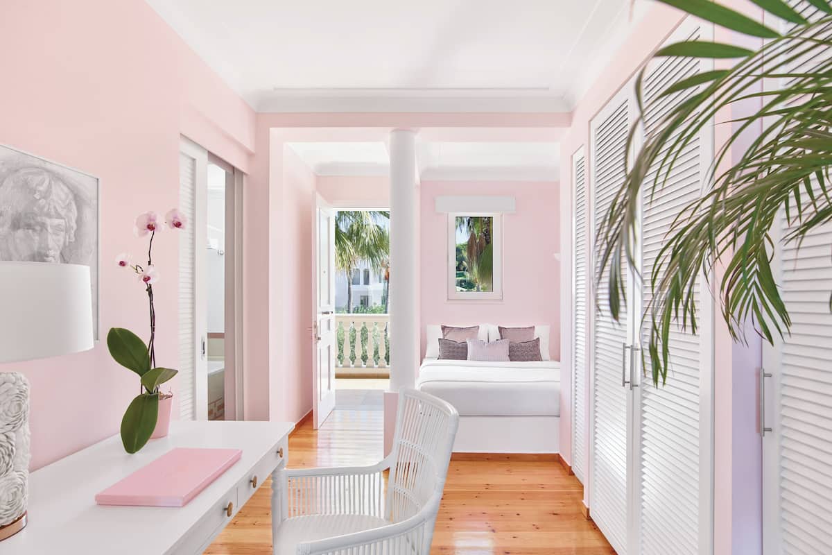A bedroom with a bed, a chair, a desk, and a window with a view of palm trees.