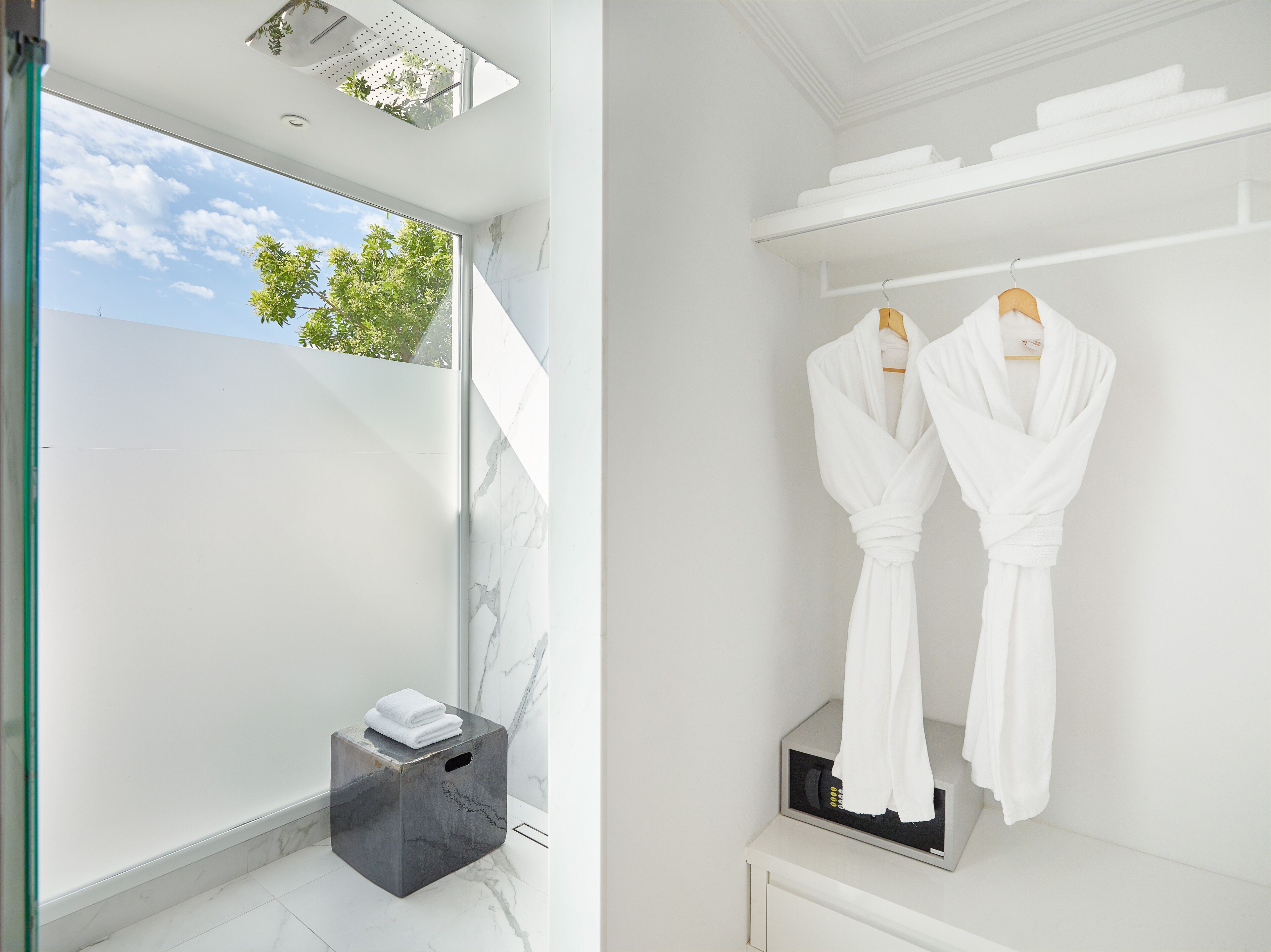 A modern bathroom with a large window, a marble vanity, and two white robes hanging on a rack.