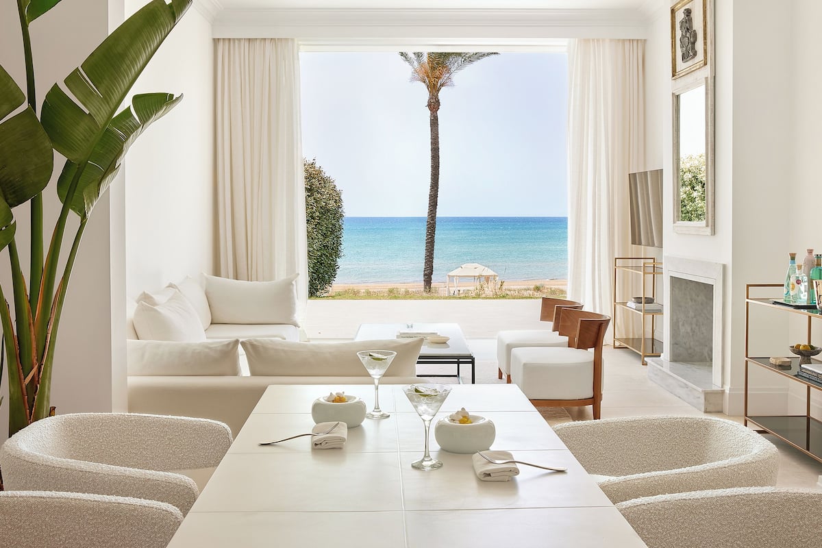 A modern hotel room with a large window overlooking a beach, a white sofa, a dining table with a martini glass, and a fireplace.