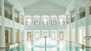 Un vaste espace intérieur abritant une piscine, avec de hauts plafonds, de grandes fenêtres et une fontaine centrale.
