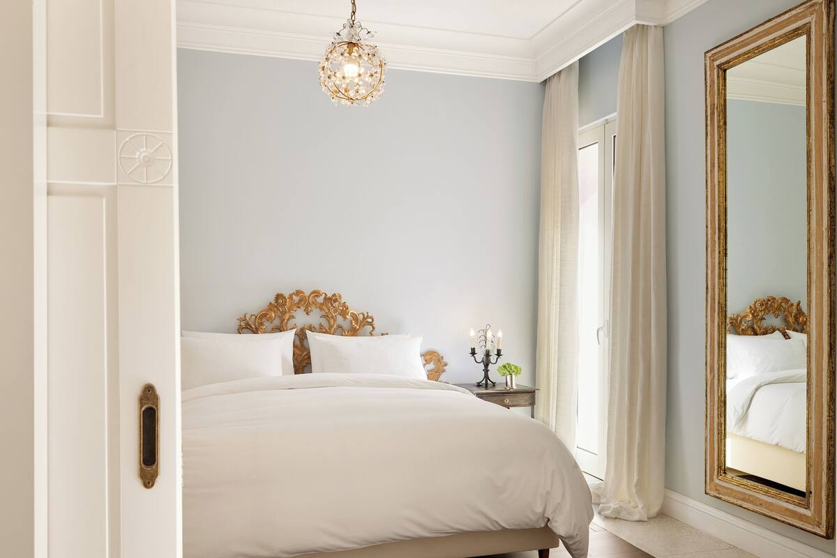 A bedroom with a large bed, a chandelier, a nightstand with a candelabra, and a mirror with a gold frame.