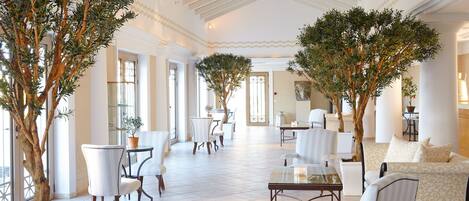 A spacious, elegantly decorated lounge area with multiple seating arrangements, large windows, and indoor plants.