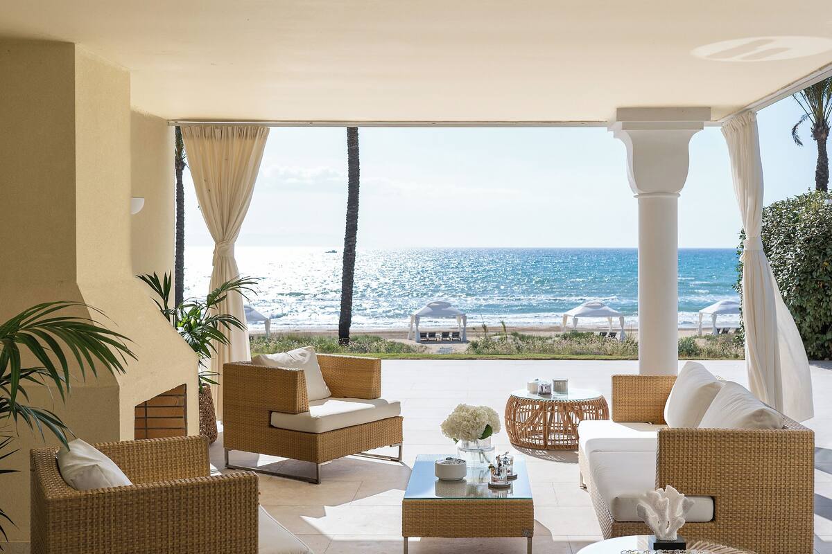 A spacious outdoor lounge area with wicker furniture, a glass coffee table, and a view of the ocean.