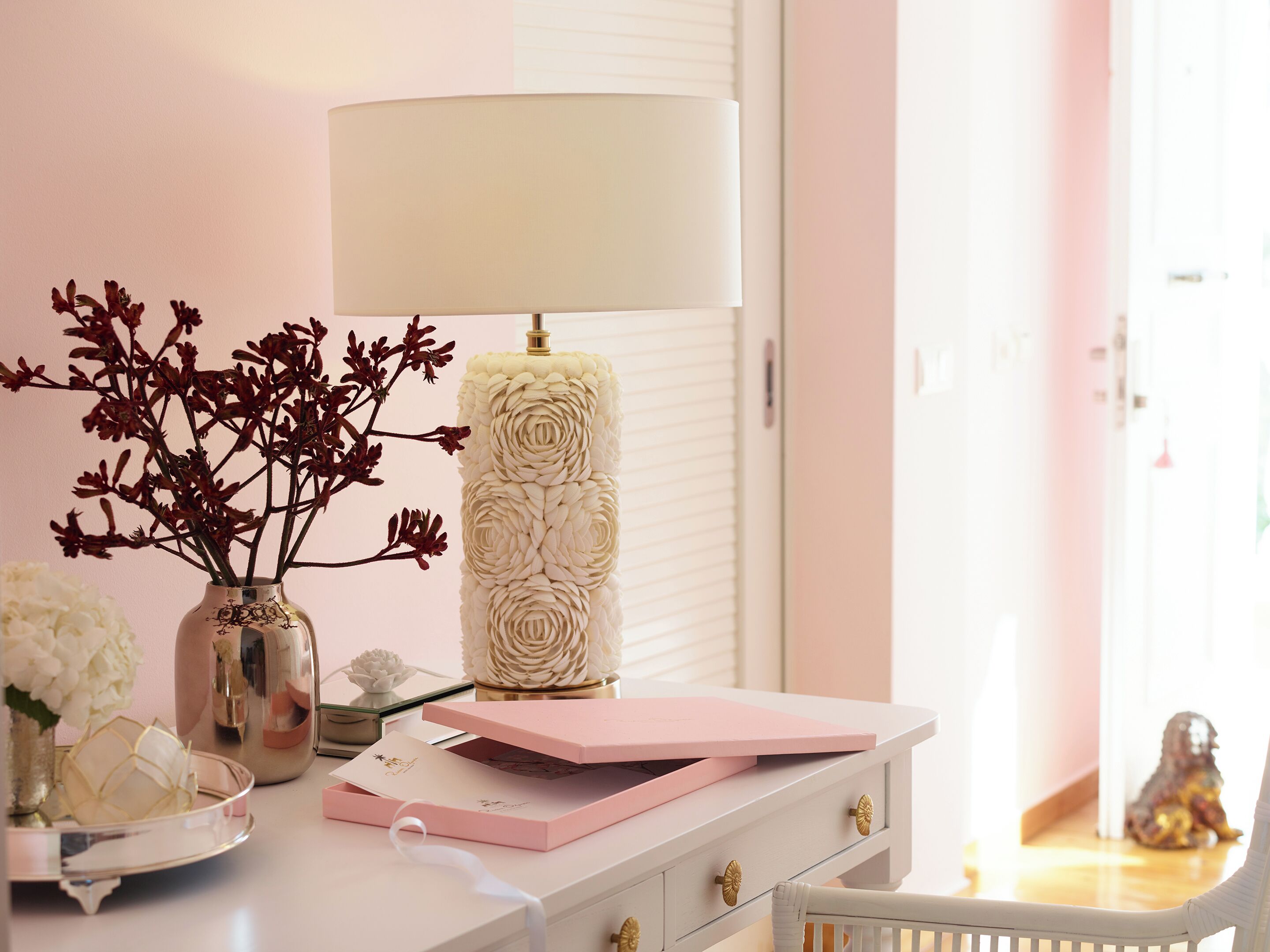 Et pænt arrangeret skrivebord med en lampe, en vase med blomster, en lille dekorativ genstand og en lyserød æske.