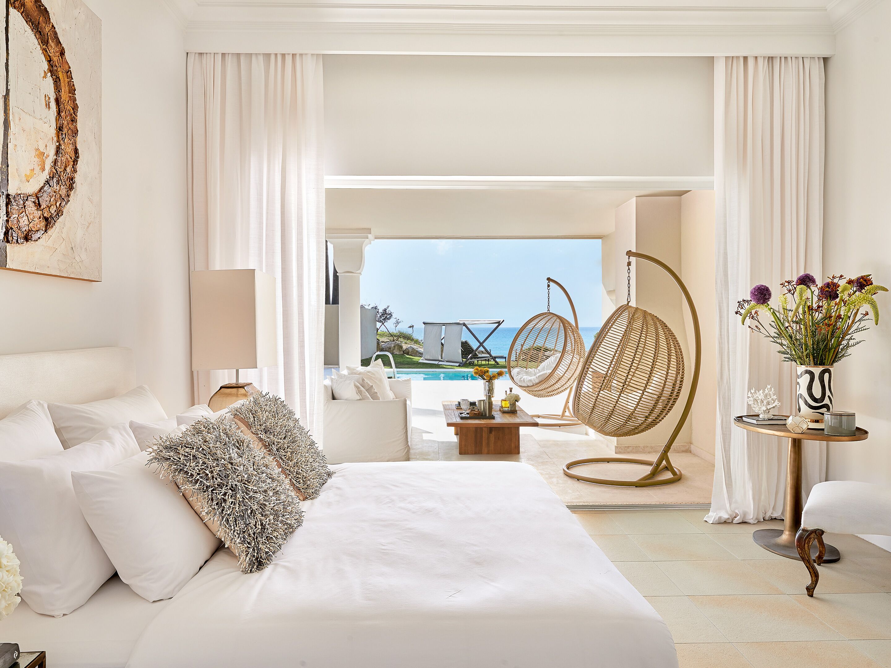 Une chambre spacieuse avec un grand lit, un fauteuil suspendu et une vue sur une piscine et l’océan.