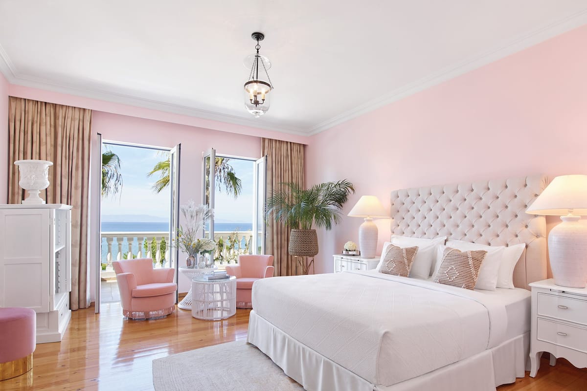A bedroom with a large bed, a nightstand, a chair, a small table, and a view of the sea through a balcony.