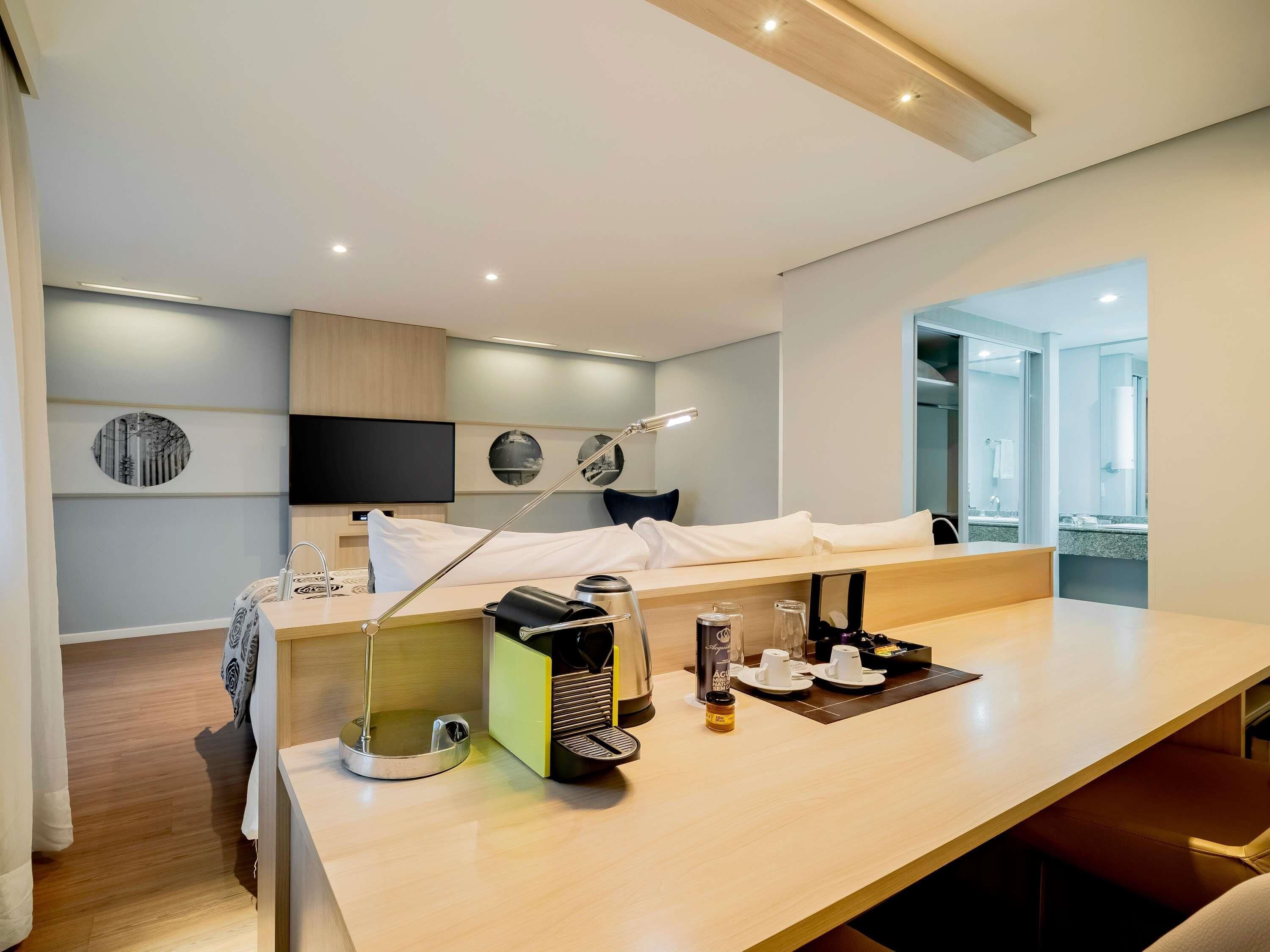 A modern hotel room with a bed, a desk with coffee-making equipment, a TV, and a bathroom visible through an open door.