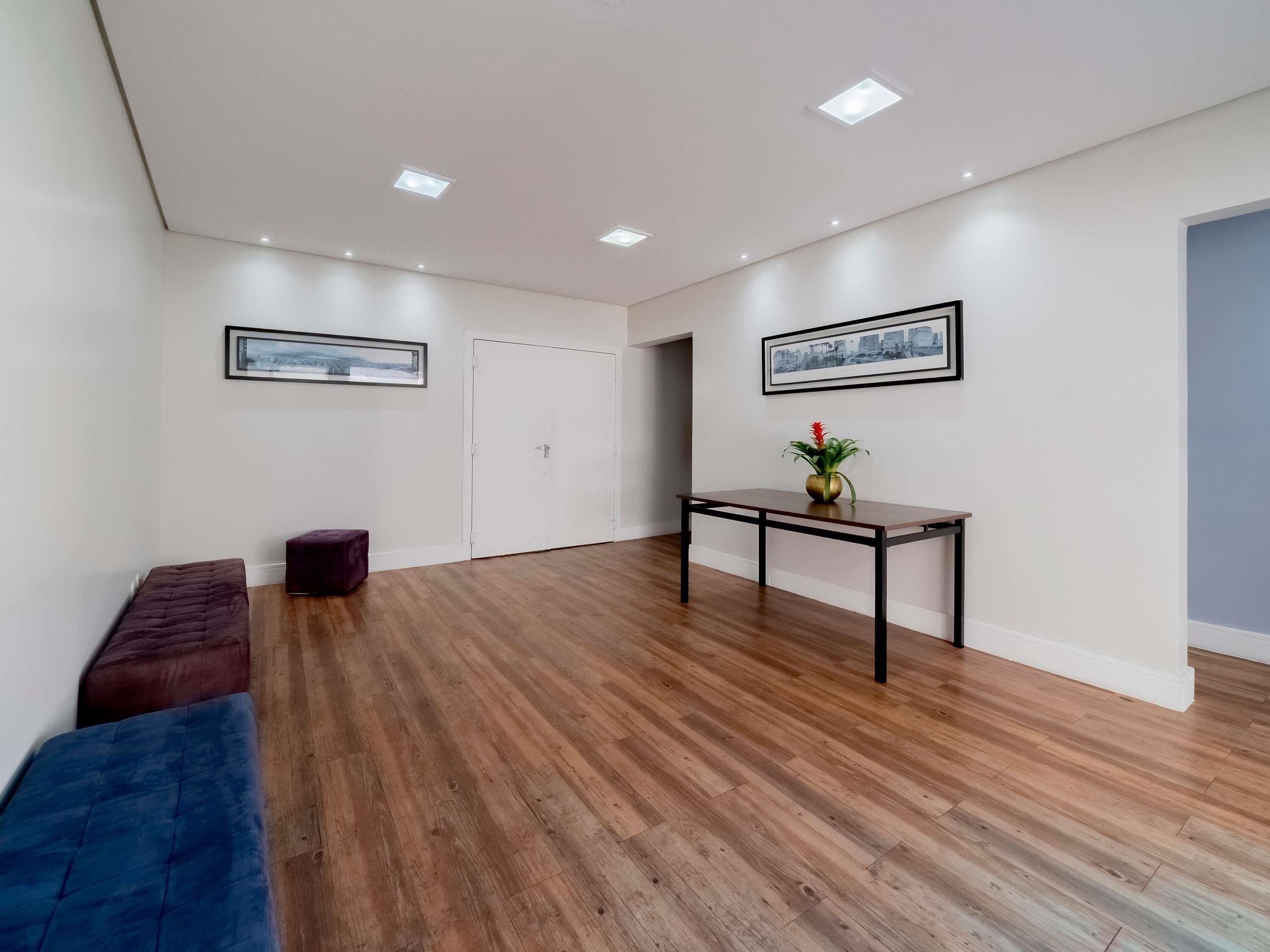 Une pièce spacieuse avec un plancher en bois, un canapé de couleur sombre, une petite table sur laquelle est posé un vase, et deux tableaux encadrés accrochés aux murs.