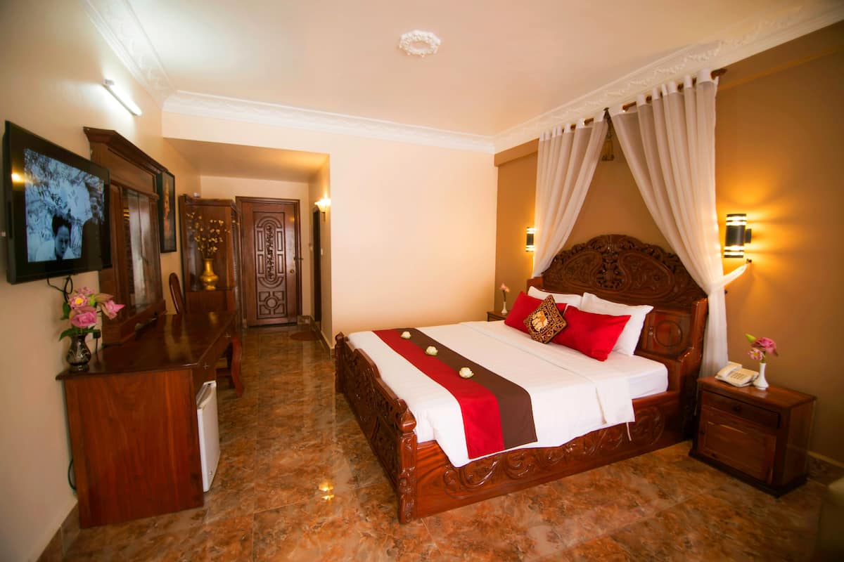 A hotel room with a wooden bed, a TV on the left, a desk with a vase of flowers, and a nightstand with a phone.