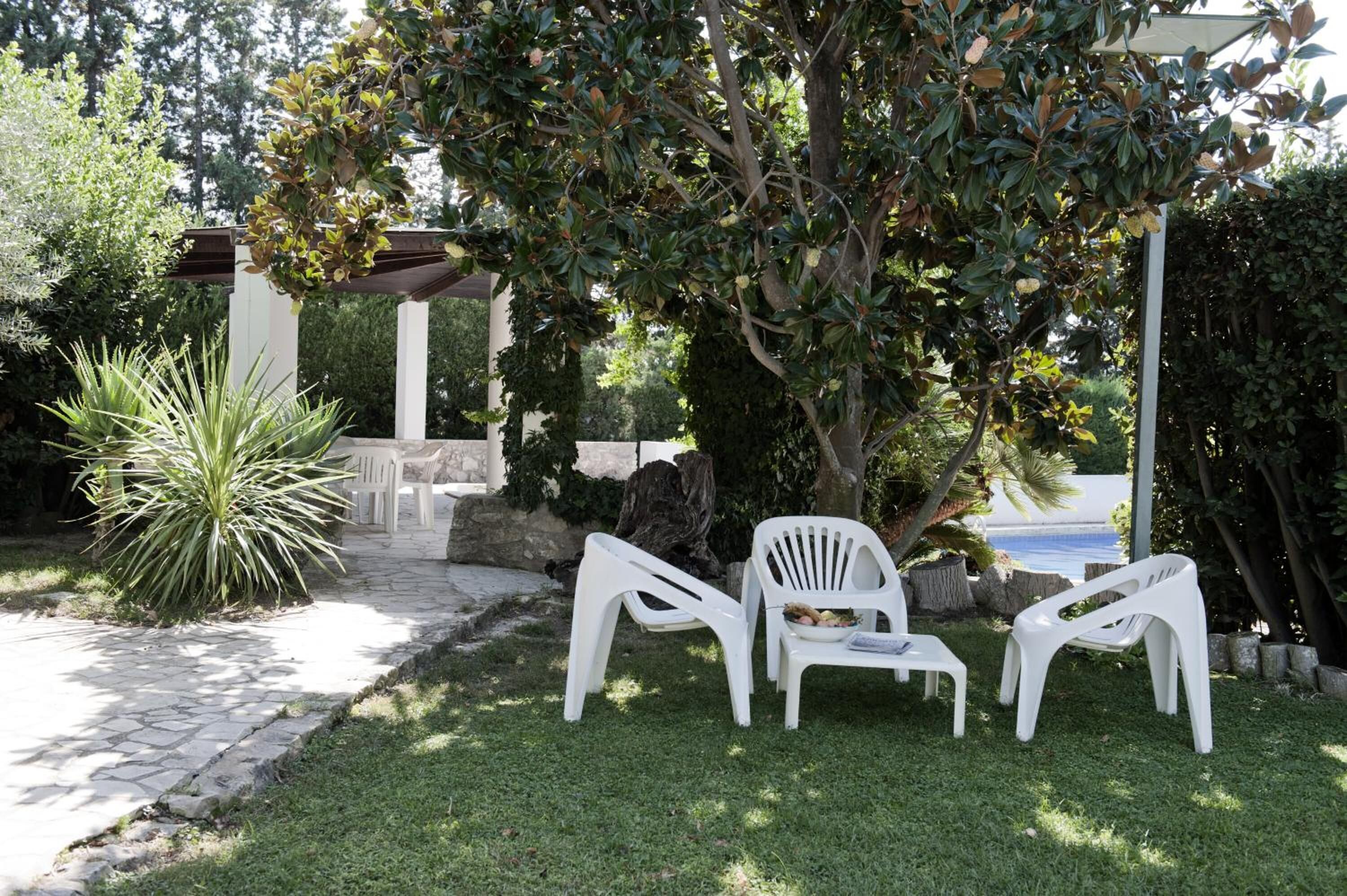 A patio area with white plastic chairs and a table, surrounded by greenery and a stone pathway.
