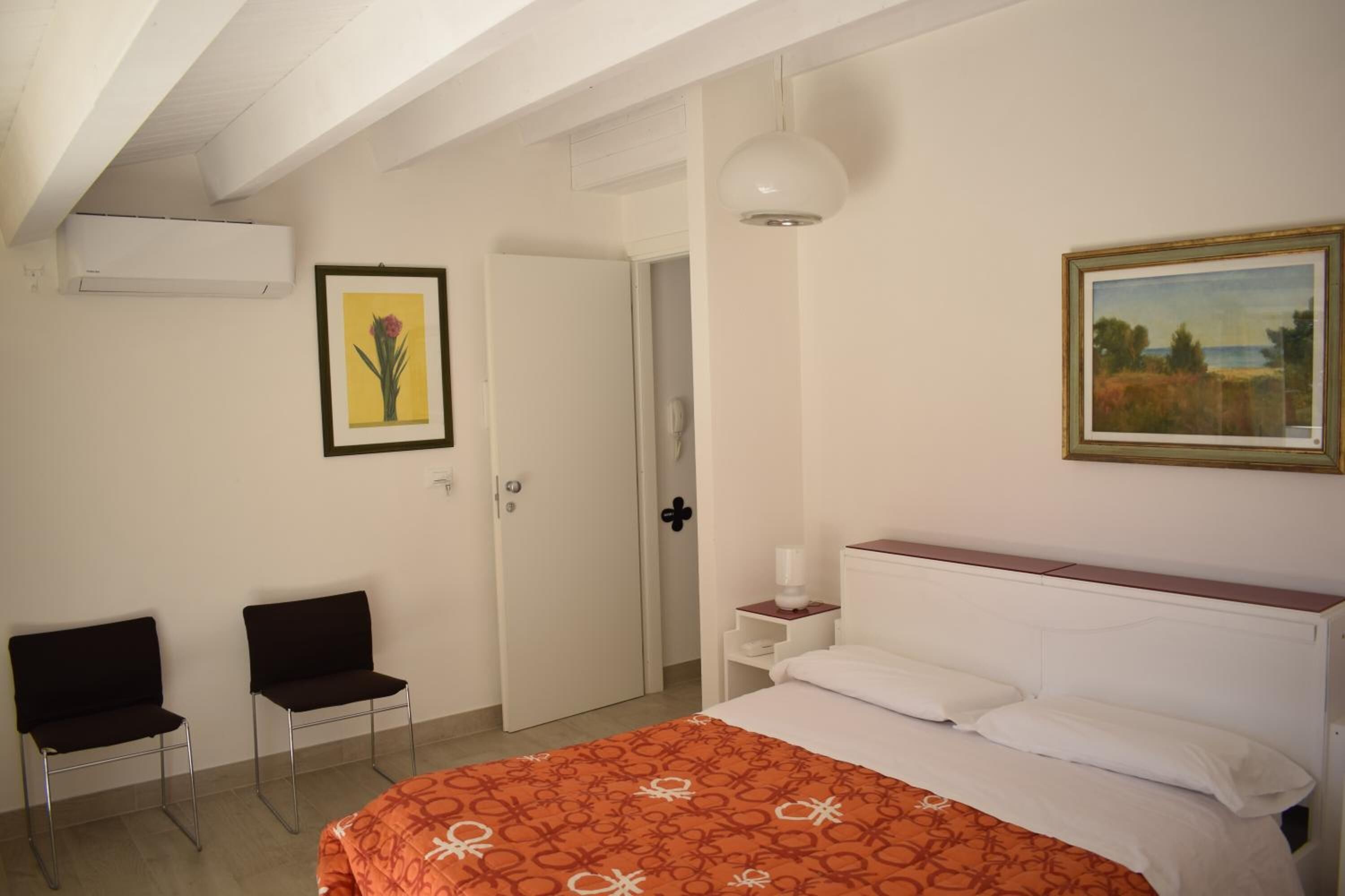 Un dormitorio con una cama, dos sillas, una mesita de noche con lámpara, una puerta y un cuadro en la pared.