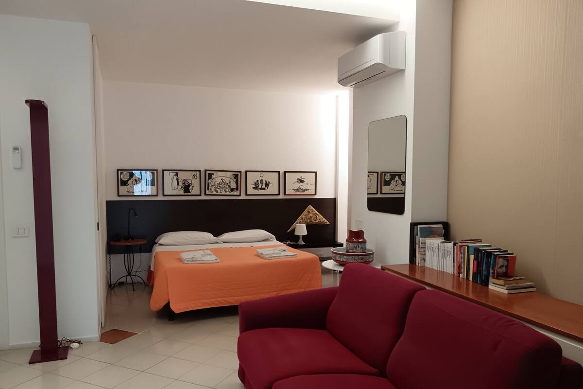 A hotel room with a bed, a red sofa, a bedside table, a lamp, and a wall with framed pictures.