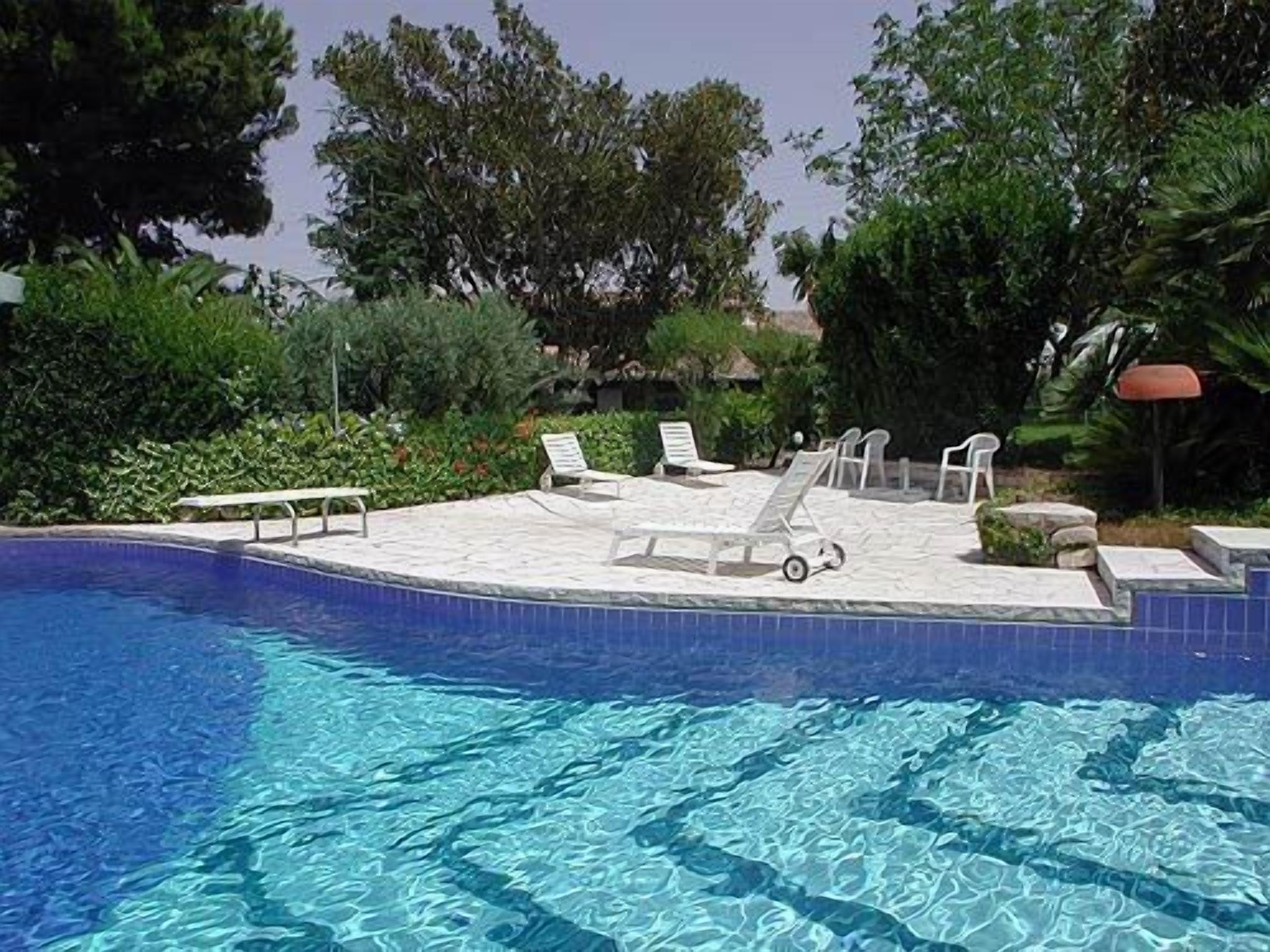 A swimming pool with clear blue water, surrounded by greenery and equipped with several lounge chairs and a small table.