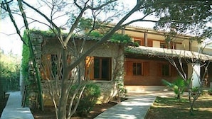 Une maison de campagne à l’aspect rustique, avec une façade en pierre et un toit de chaume, entourée de verdure et bordée d’un chemin pavé.