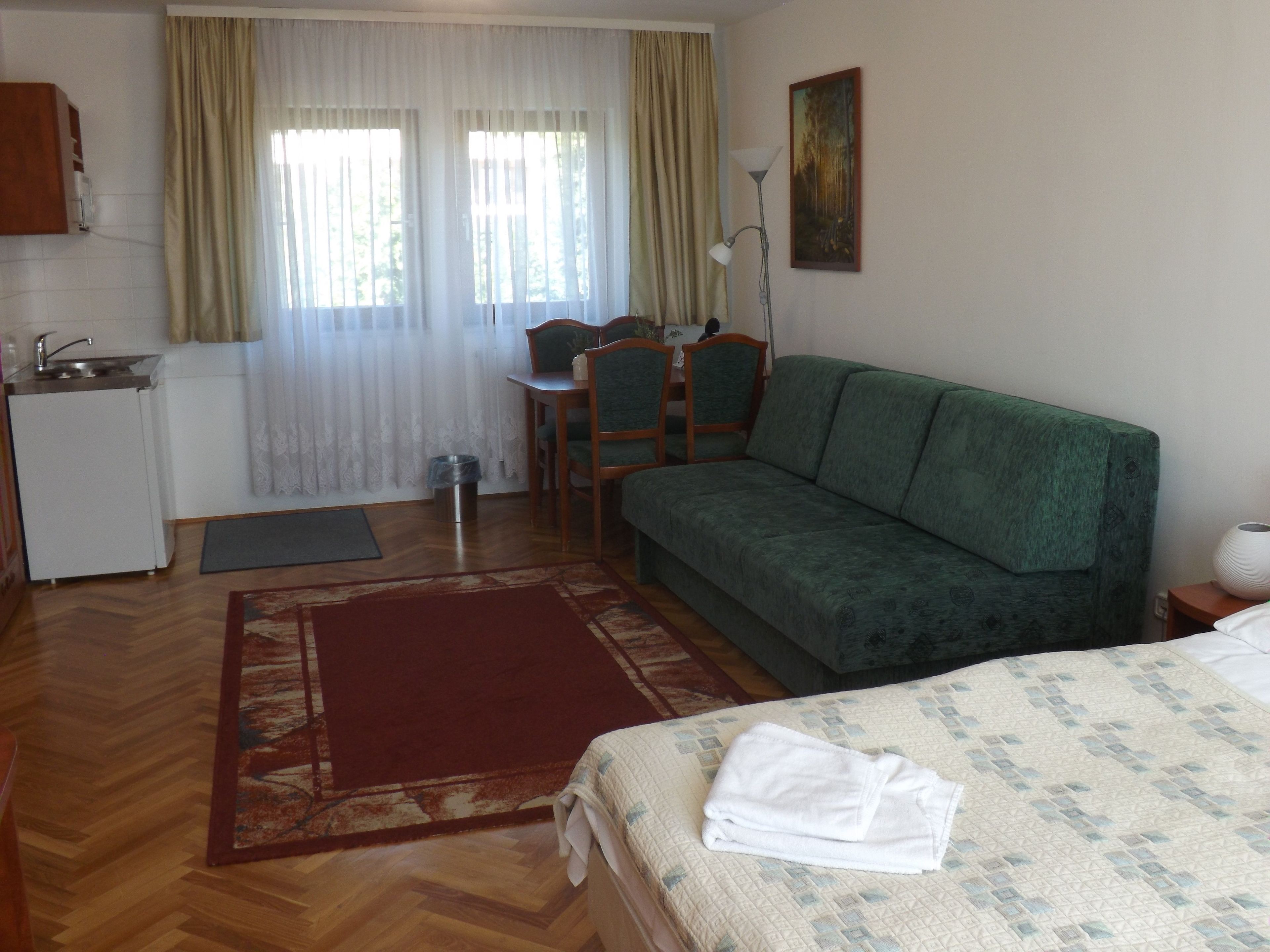 A hotel room with a bed, a green sofa, a dining table with chairs, a small kitchen area, and a painting on the wall.