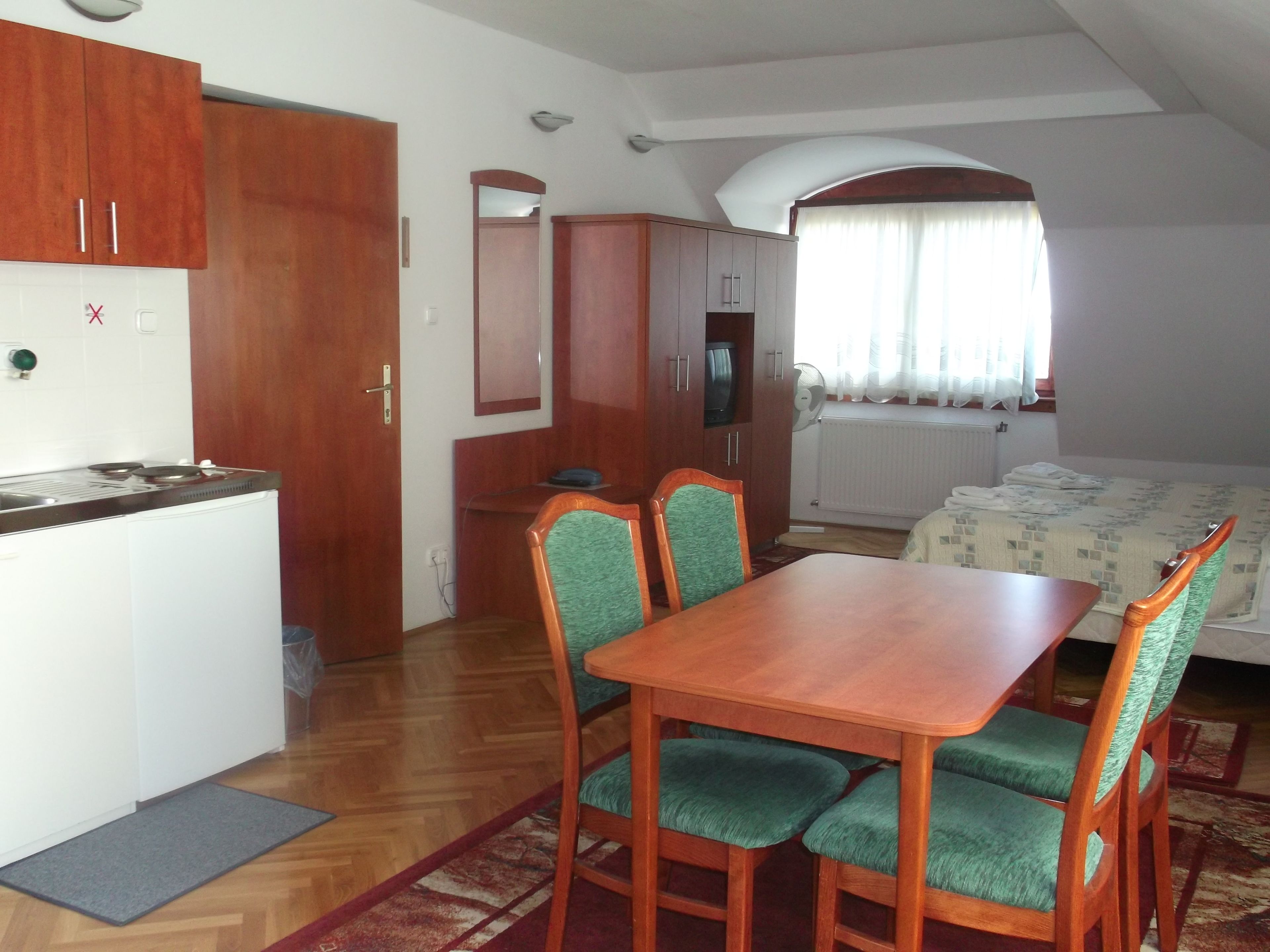 A compact room with a kitchenette, a dining area with a wooden table and green upholstered chairs, a bed with a patterned duvet, and a window with curtains.