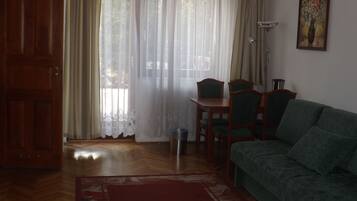 Una sala de estar con sofá verde, suelo de madera, mesa de comedor con sillas, puerta de madera y ventana con cortinas traslúcidas.