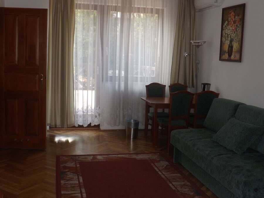 Una sala de estar con sofá verde, suelo de madera, mesa de comedor con sillas, puerta de madera y ventana con cortinas traslúcidas.