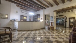 Un hall d’hôtel avec un sol à carreaux, des poutres en bois au plafond, un comptoir d’accueil et une vue sur l’extérieur par le biais d’une arche.