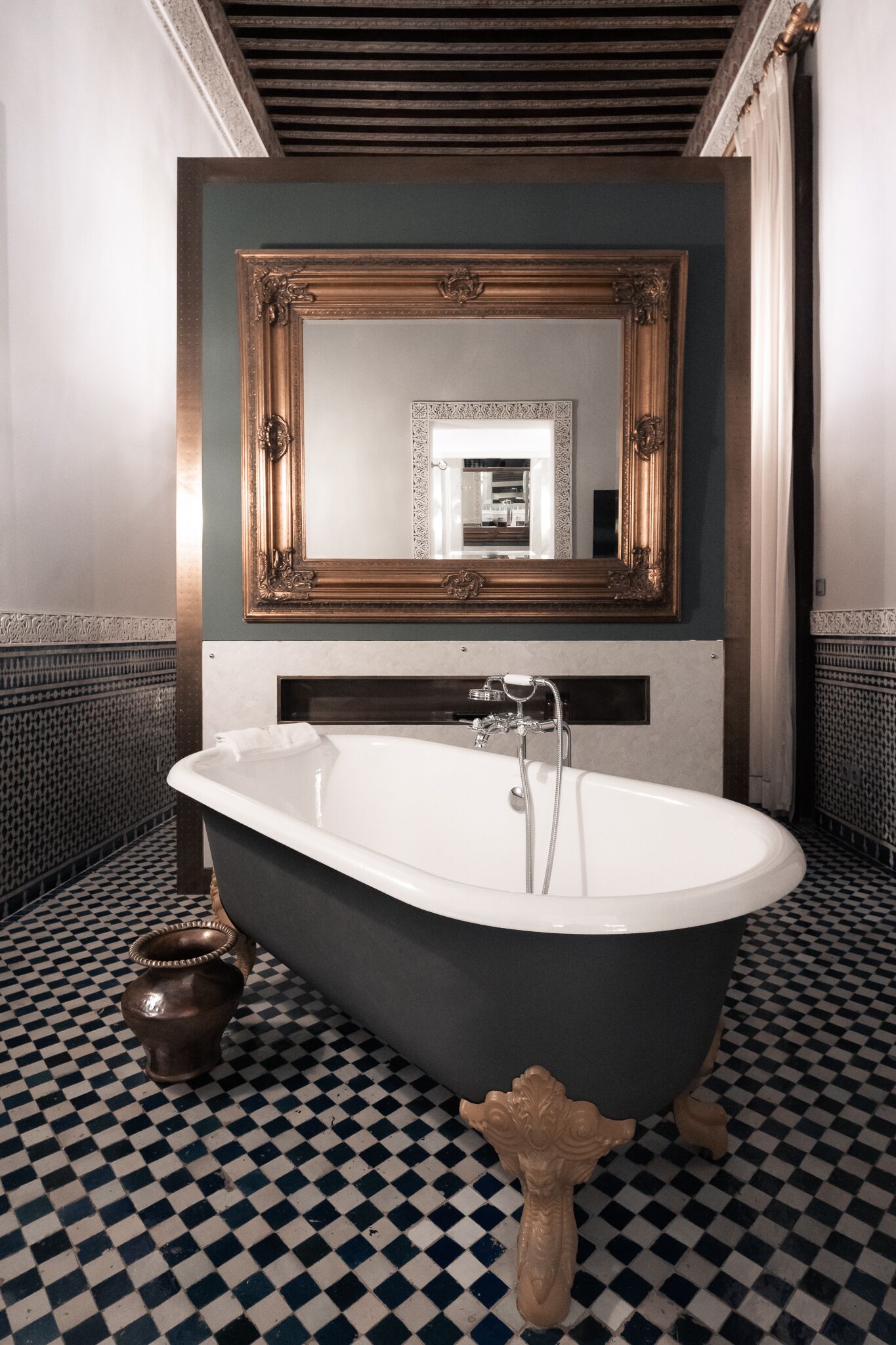 Ein Badezimmer mit Schachbrettboden, einer freistehenden Badewanne, einem großen Spiegel mit aufwendigem Rahmen und einer kleinen Dekorationsvase.