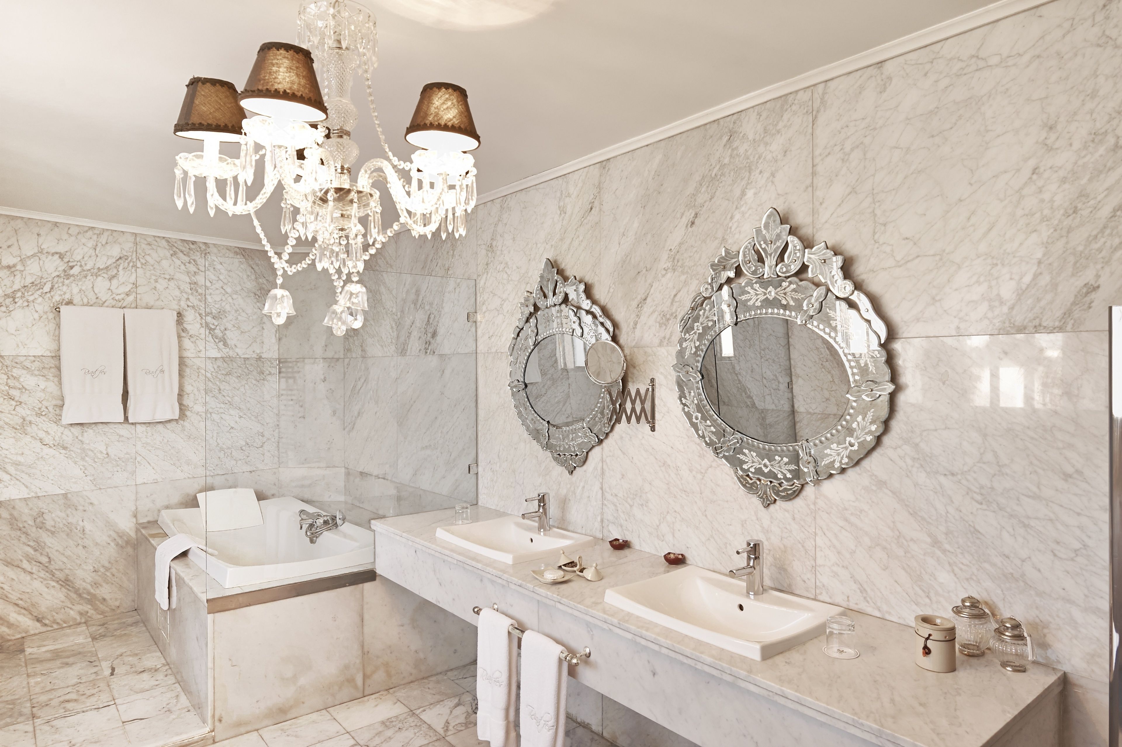 Une salle de bain luxueuse avec un grand mur de marbre, un somptueux lustre et des miroirs richement décorés.