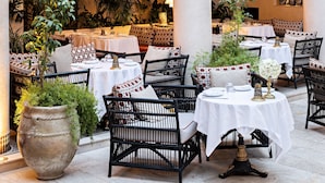 Un espace extérieur aménagé pour les repas, avec des tables blanches et des chaises en osier noir, entouré d’arches décoratives et de plantes en pot.