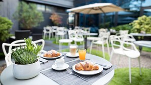 An outdoor dining area with white chairs and tables, a potted plant, croissants, a cup of coffee, and a glass of orange juice.