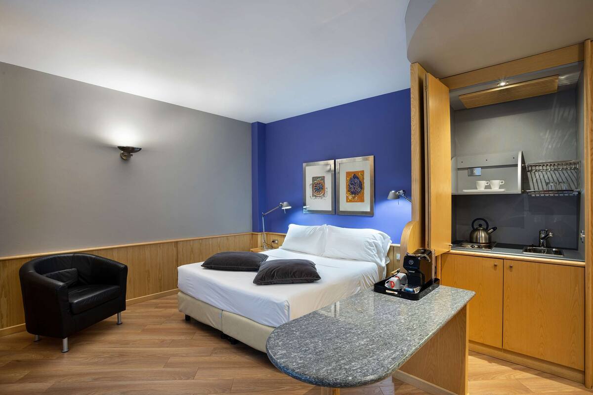 A compact hotel room with a bed, a small seating area, a kitchenette, and a wall-mounted light fixture.
